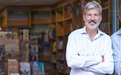 Middle aged man with gray beard smiling while leaning against the front door of his bookstore